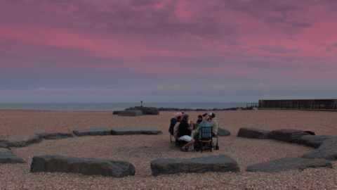 A group of people seated around a stone circle on a beach, under a vibrant pink and purple sky at dusk.