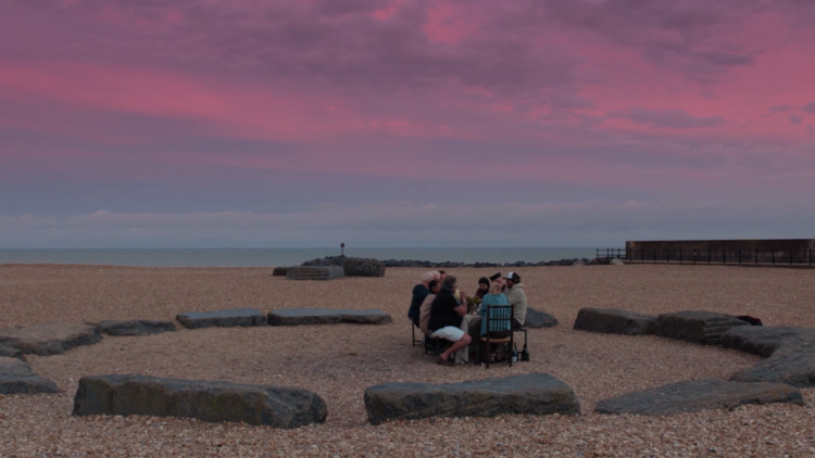 A group of people seated around a stone circle on a beach, under a vibrant pink and purple sky at dusk.