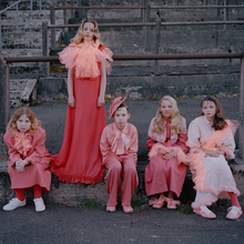 A group of children in matching pink and coral outfits stands on a stone ledge, posing against a textured backdrop of steps.