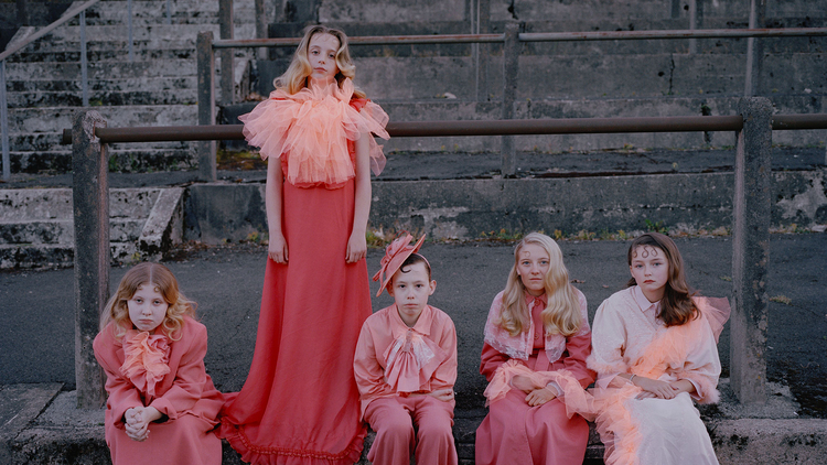 A group of children in matching pink and coral outfits stands on a stone ledge, posing against a textured backdrop of steps.