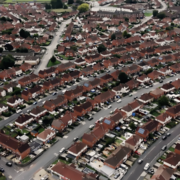 Aerial view of a suburban neighborhood featuring rows of red-roofed houses, winding roads, and greenery throughout the area.