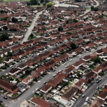 Aerial view of a suburban neighborhood featuring rows of red-roofed houses, winding roads, and greenery throughout the area.