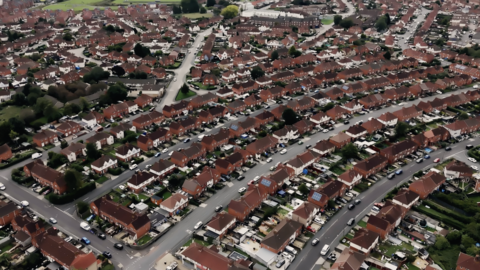 Aerial view of a suburban neighborhood featuring rows of red-roofed houses, winding roads, and greenery throughout the area.