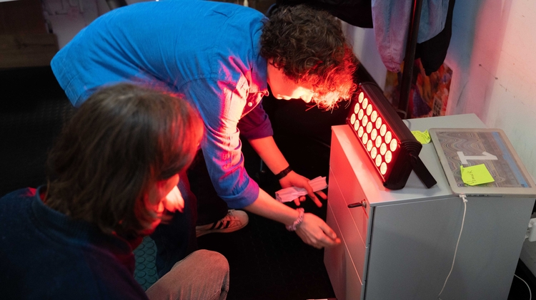 Two people interact with a glowing red light source in a dim room. The atmosphere suggests curiosity and focus, with scattered objects around.