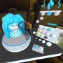 A screenshot from Taru Muhonen's app, Spira - the image depicts a digital render of the app in use; in the centre of the shot, a transparent hand, controlled by the user, fixes a bust with a white face and blue hair. To the right of the bust is a white controller with multi coloured buttons. The bust and controller sits on a black tabletop. 