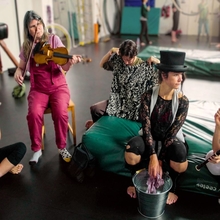 A group of circus performers, all white women, sit in a semi circle in a rehearsal space with a black floor. Some sit on the floor, while others sit on green cushions or chairs. They are in discussion, while two performers on the left play instruments - one, on the far left, plays the flute, while the other to her right plays a fiddle. 