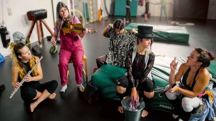 A group of circus performers, all white women, sit in a semi circle in a rehearsal space with a black floor. Some sit on the floor, while others sit on green cushions or chairs. They are in discussion, while two performers on the left play instruments - one, on the far left, plays the flute, while the other to her right plays a fiddle. 
