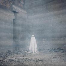 A ghostly figure in a white sheet stands amid a foggy, industrial construction site, with concrete walls and scattered debris.