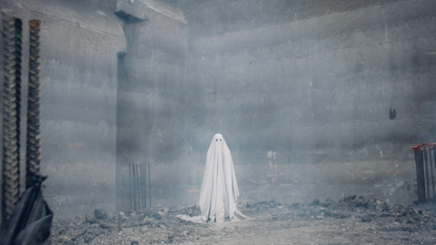 A ghostly figure in a white sheet stands amid a foggy, industrial construction site, with concrete walls and scattered debris.