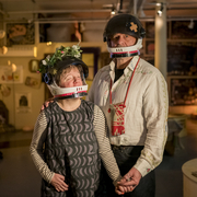 Andrew and Eden Kotting are in a warmly lit room wear black helmets with flower and bandage decorations. They hold hands, conveying a sense of connection and warmth.
