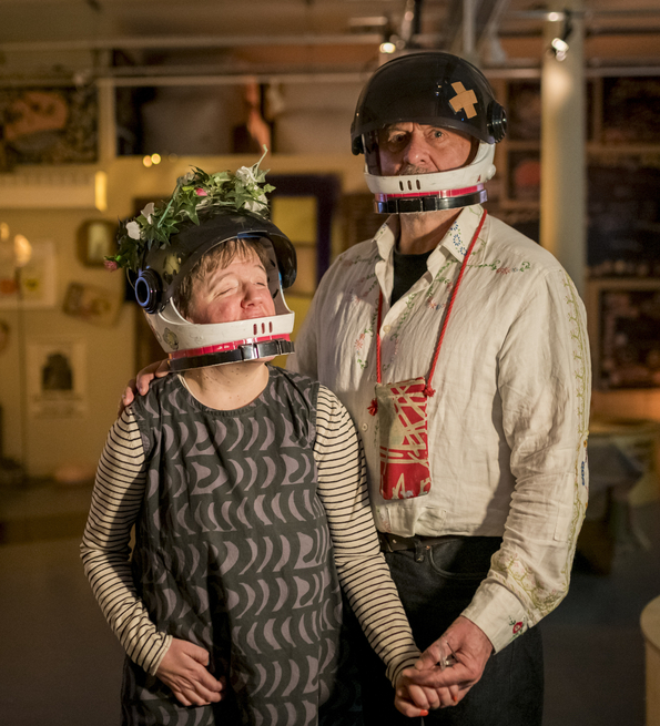 Andrew and Eden Kotting are in a warmly lit room wear black helmets with flower and bandage decorations. They hold hands, conveying a sense of connection and warmth.