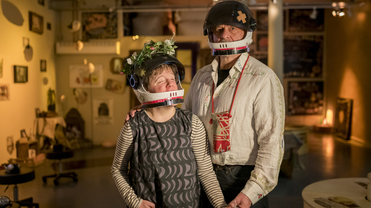 Andrew and Eden Kotting are in a warmly lit room wear black helmets with flower and bandage decorations. They hold hands, conveying a sense of connection and warmth.