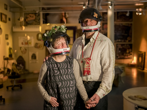 Andrew and Eden Kotting are in a warmly lit room wear black helmets with flower and bandage decorations. They hold hands, conveying a sense of connection and warmth.