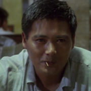 A person sits in a dimly lit restaurant, focused on a conversation, with another person partially visible in the background reading a newspaper.