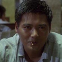 A person sits in a dimly lit restaurant, focused on a conversation, with another person partially visible in the background reading a newspaper.