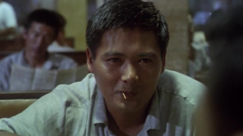 A person sits in a dimly lit restaurant, focused on a conversation, with another person partially visible in the background reading a newspaper.