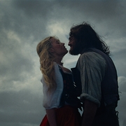 A couple stands close, sharing a tense embrace against a dramatic cloudy sky, evoking a sense of romance and adventure.