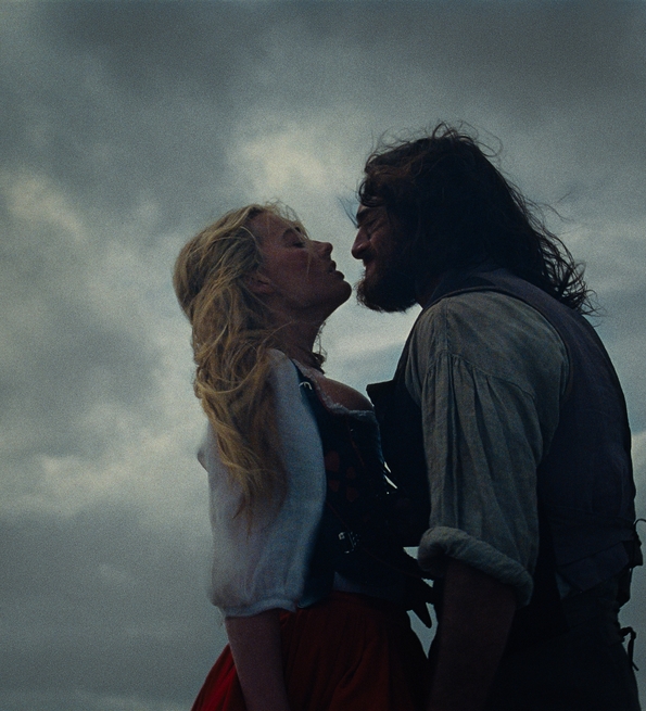 A couple stands close, sharing a tense embrace against a dramatic cloudy sky, evoking a sense of romance and adventure.