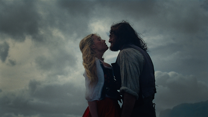 A couple stands close, sharing a tense embrace against a dramatic cloudy sky, evoking a sense of romance and adventure.