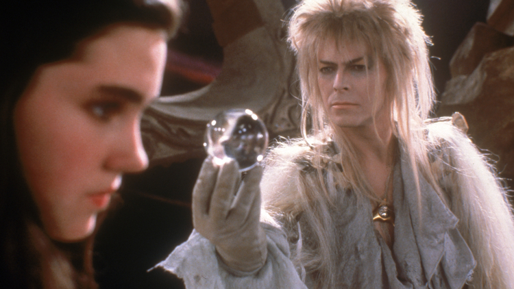 A character with flowing hair holds a crystal ball, surrounded by a mystical setting with rocks and shadows.