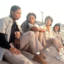 A group of four women sits on a rooftop, casually dressed, enjoying drinks and music from a boombox, with a city backdrop.