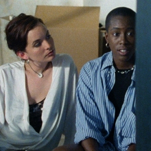 Two people sit together in a casual setting, one holding a glass of red wine, surrounded by minimal decor and a sense of intimacy.