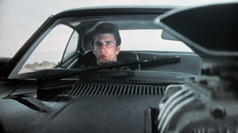 A close-up view from inside a classic car, showing the dashboard and engine details, with a focus on the driver.