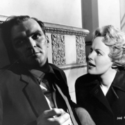 A man and a woman sit closely together in a dimly lit space, both dressed in formal attire, with ornate decor in the background.