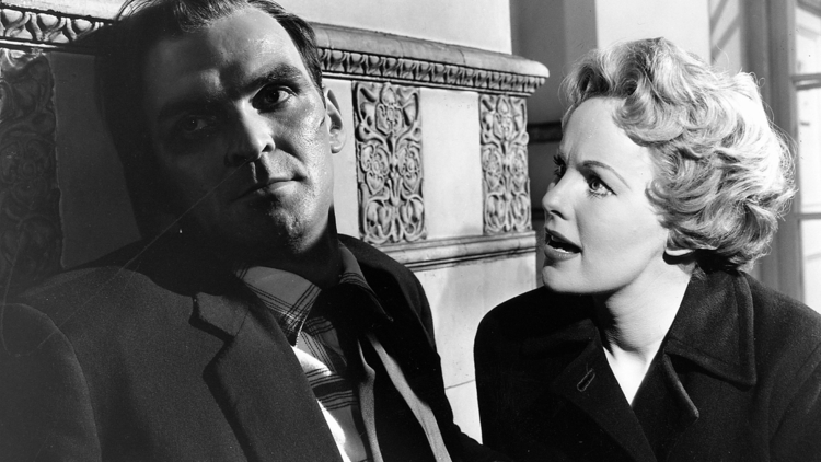 A man and a woman sit closely together in a dimly lit space, both dressed in formal attire, with ornate decor in the background.