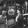 A black-and-white image of two men talking near a truck marked "SOC," with workers loading equipment in the background.