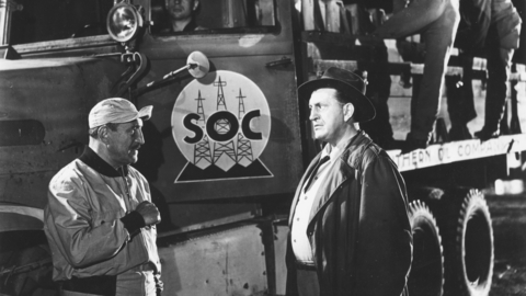 A black-and-white image of two men talking near a truck marked "SOC," with workers loading equipment in the background.