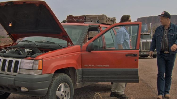 A red Jeep Grand Cherokee with its hood raised, parked on a dirt road amid rocky terrain, while two people converse nearby.