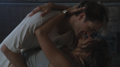 A couple embraces intimately, their faces close, surrounded by a softly lit, rustic indoor setting.