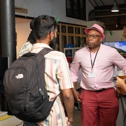 A group of four people engage in conversation in an office setting. A person in a pink hat and shirt stands at the center. Casual and friendly atmosphere.