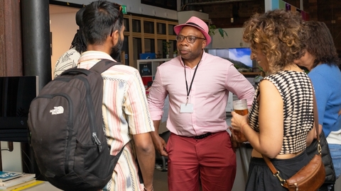 A group of four people engage in conversation in an office setting. A person in a pink hat and shirt stands at the center. Casual and friendly atmosphere.