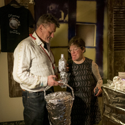 Andrew and Eden Kötting are smiling warmly stand next to a table covered in foil-wrapped items, suggesting a cozy, quirky atmosphere in a creatively decorated room.