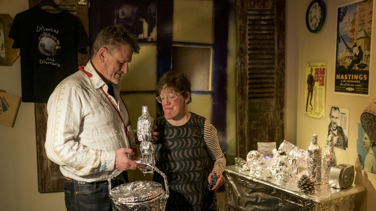 Andrew and Eden Kötting are smiling warmly stand next to a table covered in foil-wrapped items, suggesting a cozy, quirky atmosphere in a creatively decorated room.