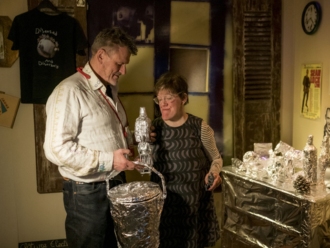 Andrew and Eden Kötting are smiling warmly stand next to a table covered in foil-wrapped items, suggesting a cozy, quirky atmosphere in a creatively decorated room.