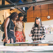 Four people gather around a table examining artwork in a well-lit room. The expressions convey curiosity and engagement, suggesting a collaborative setting.