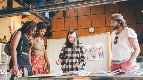 Four people gather around a table examining artwork in a well-lit room. The expressions convey curiosity and engagement, suggesting a collaborative setting.