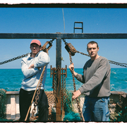 Two individuals stand on a fishing boat, with fishing nets and equipment visible in the background, under a clear blue sky.