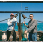 Two individuals stand on a fishing boat, with fishing nets and equipment visible in the background, under a clear blue sky.