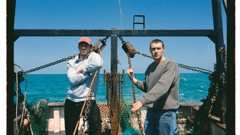 Two individuals stand on a fishing boat, with fishing nets and equipment visible in the background, under a clear blue sky.