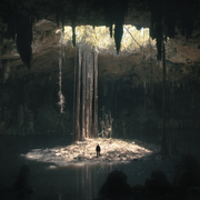 A lone figure stands on a small island in a dark cavern, surrounded by water, with sunlight streaming through a large opening above.