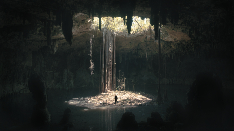 A lone figure stands on a small island in a dark cavern, surrounded by water, with sunlight streaming through a large opening above.