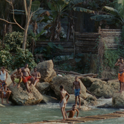 A group of people play by a river, perched on rocks surrounded by lush greenery and a rustic wooden background.