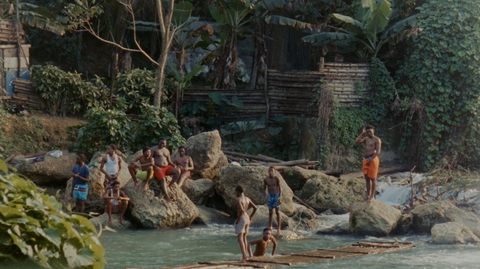 A group of people play by a river, perched on rocks surrounded by lush greenery and a rustic wooden background.