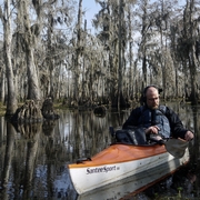 Man in an orange kayak paddles through a calm swamp with tall cypress trees draped in moss. He wears a life jacket and headphones, conveying focus.