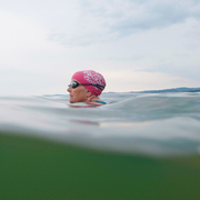 Swimmer wearing a pink swim cap in open water under a cloudy sky with distant shoreline in the background.