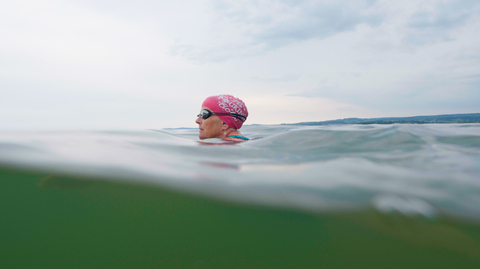 Swimmer wearing a pink swim cap in open water under a cloudy sky with distant shoreline in the background.